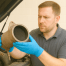 Mechanic in blue gloves inspecting a diesel particulate filter in a bright, sunlit workshop.