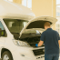 Mechanic in blue gloves inspecting the engine of a white motorhome, back facing the camera in a bright workshop.
